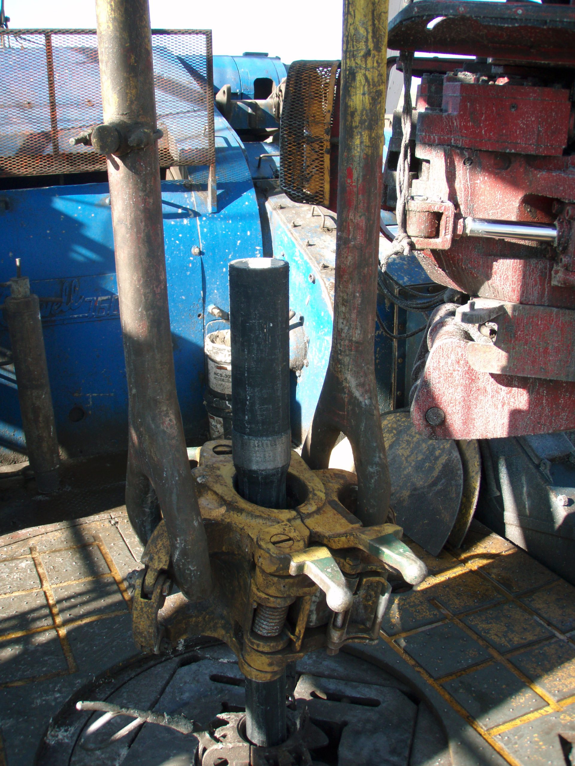 A drillstring joint in an elevator above the rotary table, beside rig-floor handling equipment.